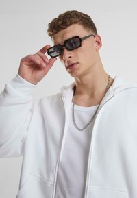 Čierne obdĺžnikové slnečné okuliare, biele mikina na zips, rebrovaný biely tielko a strieborný náhrdelník. Model umiestnený pod uhlom.