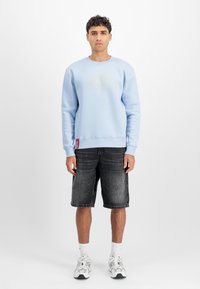 Világoskék kapucnis pulóver középső arany logóval, fekete farmertortával és fehér sportcipővel párosítva. A pulóver bordás mandzsettákkal rendelkezik.