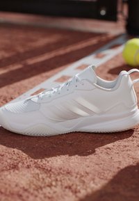 Scarpa sportiva bianca su un campo da tennis in terra rossa vicino a una linea bianca di confine con una palla da tennis gialla sullo sfondo.