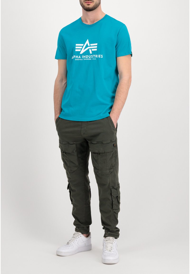 Tyrkysové bavlnené tričko s bielym logom "Alpha Industries"; olivovozelené cargo nohavice s viacerými vreckami; biele tenisky.