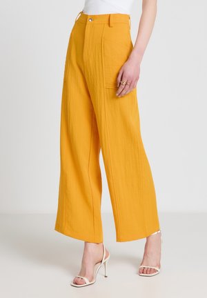 Femme portant un pantalon large jaune moutarde avec une poche plaquée à l'avant et des sandales blanches à talons hauts, debout sur un fond blanc.