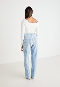 Kvinna som står vänd bort, klädd i en asymmetrisk vit långärmad topp, ljusblå högmidjade jeans och vita spetsiga klackskor.