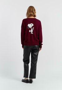Personne avec des cheveux mi-longs portant un pull bordeaux avec un graphique Snoopy, un pantalon en cuir noir, des chaussettes beiges et des mocassins noirs, de dos.