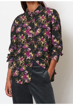 Femme portant une chemise noire à manches longues à boutons avec des fleurs violettes, roses et jaunes, associée à un pantalon en velours côtelé gris foncé.