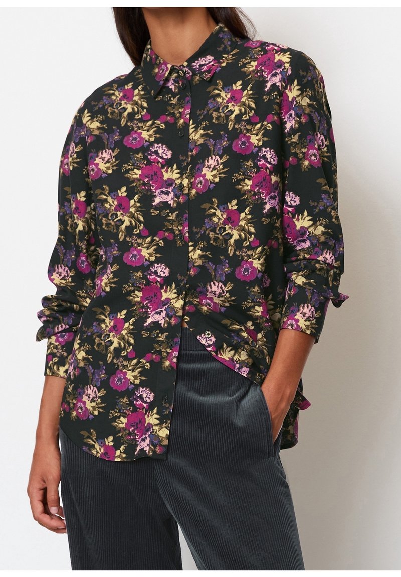 Femme portant une chemise noire à manches longues à boutons avec des fleurs violettes, roses et jaunes, associée à un pantalon en velours côtelé gris foncé.
