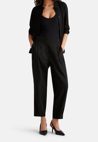 Svart skjorta med knappar ovanpå en svart bodysuit, kombinerad med höga, plisserade svarta byxor och spetsiga svarta klackskor.