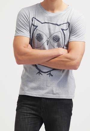 Mężczyzna w szarej koszulce z dużym czarnym szkicem sowy, z założonymi skrzyżowanymi ramionami, w czarnych dżinsach na jednolitym tle.