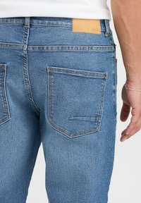Jeans de mezclilla azul con una textura suave, que presentan dos bolsillos traseros, costuras en contraste y un parche de cuero marrón claro en la cintura.
