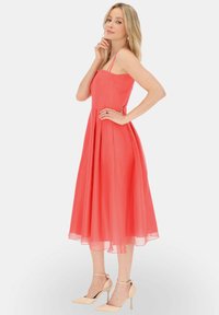 Koralowa sukienka typu A z cienkimi ramiączkami, plisowanym dołem i długością do kolan. Modelka w cielistych szpilkach stoi na białym tle.