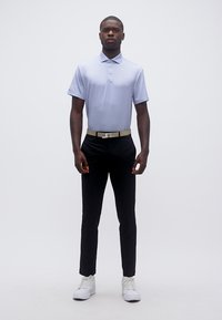 Άνδρας που στέκεται φορώντας ανοιχτό μπλε πόλο πουκάμισο, μαύρο παντελόνι, μπεζ ζώνη και λευκά αθλητικά παπούτσια μπροστά σε απλό λευκό φόντο.