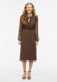 Brun prikkete kjole med lange, gjennomsiktige ermer, knyting i halsen og innsvingt midje. Kjolen går under knærne, kombinert med svarte hæler med ankelrem.