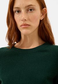 Dunkelgrüner Strickpullover mit rundem Hals und subtiler Textur. Der Stoff wirkt weich und hat eine leicht lockere Passform an den Schultern.