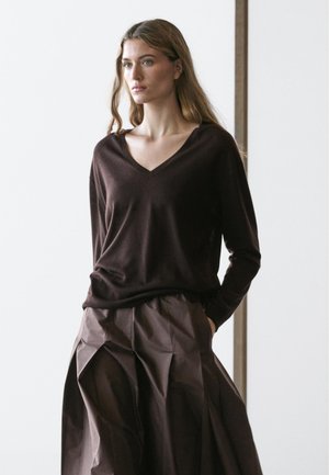 Donna con capelli lunghi che indossa un maglione scollo a V marrone scuro e una gonna plissettata marrone, in piedi con le mani nelle tasche.