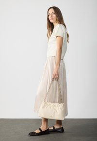 Světle béžový top s krátkými rukávy, plisovaná lesklá stříbrná midi sukně, černé ploché boty a krémová kabelka s nařaseným designem a řetízkovým popruhem.