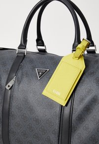 Sac à main synthétique gris avec des accents en cuir noir, arborant un logo Guess triangulaire et une étiquette de bagage jaune vif. Texture lisse, fermeture à glissière.