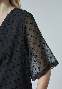 Sort bluse med gennemsigtige korte ærmer, der har strukturerede polkaprikker på et gittermønster, båret af en person med lys hud og blondt hår.