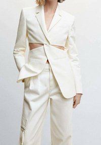 Femme portant un blazer blanc court avec une découpe au niveau de la taille et un pantalon taille haute assorti avec un détail de fermeture éclair sur la poche latérale.