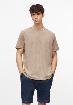 Jeune homme debout les mains dans les poches, portant un t-shirt beige à manches courtes et un short bleu foncé sur fond blanc.