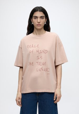 Giovane donna con lunghi capelli scuri indossa una maglietta oversize rosa chiaro con la scritta "Peace of mind is the true luxury" e jeans blu.