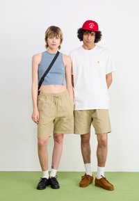 Two individuals stand side by side. The person on the left wears a light blue cropped tank, beige shorts, and black shoes. The person on the right has a white t-shirt, beige shorts, and brown shoes.