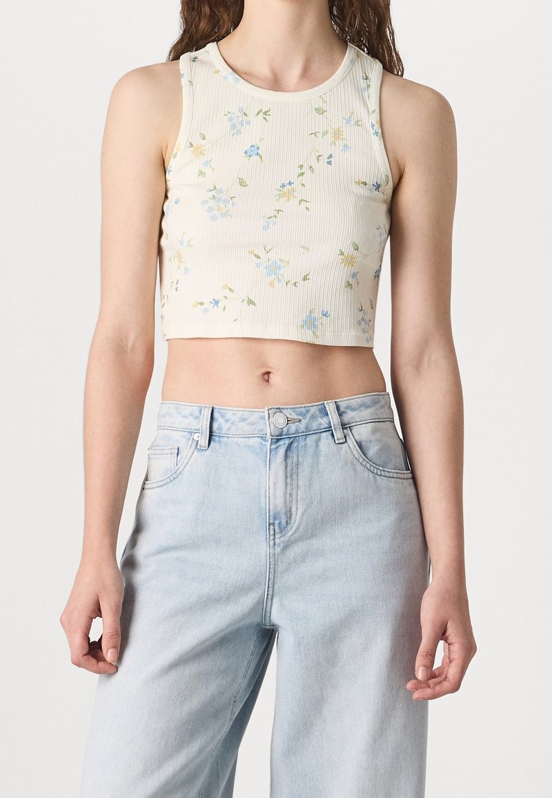 Femme portant un crop top crème sans manches avec un petit imprimé floral bleu et un jean taille haute bleu clair.