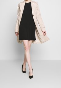 Beige trenchcoat mustana mekko, jossa on pitkät hihat ja muokattu malli. Mallilla on mustat teräväkärkiset korkokengät.