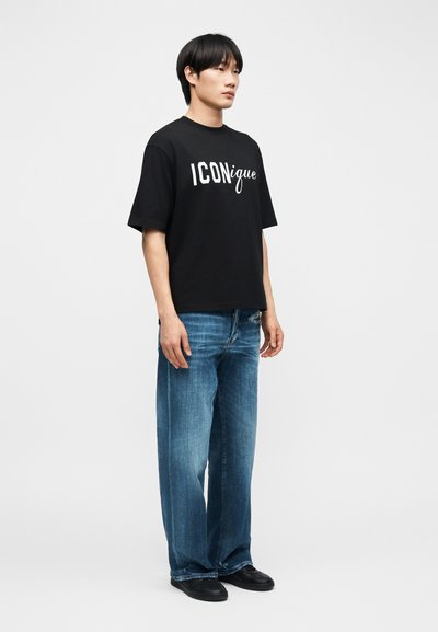 Νέος άνδρας στέκεται φορώντας μαύρο T-shirt με το λογότυπο "ICONique", φαρδιά μπλε τζιν και μαύρα παπούτσια, με απλό φωτεινό φόντο.