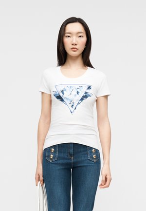 Mujer joven con camiseta blanca con gráfico triangular azul y vaqueros azul oscuro con botones dorados, sosteniendo una bolsa blanca con rayas.