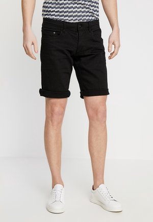 Mann trägt hochgekrempelte schwarze Jeans-Shorts, weiße Turnschuhe und ein gestreiftes Kurzarmhemd, steht vor weißem Hintergrund.