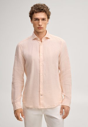 Young man wearing a light peach long-sleeve button-up shirt and white pants, standing against a neutral background.