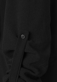 Manica di blusa nera con una sottile texture, caratterizzata da una fascia con bottone e un design di tessuto arricciato, che mette in evidenza dettagli minimalisti.