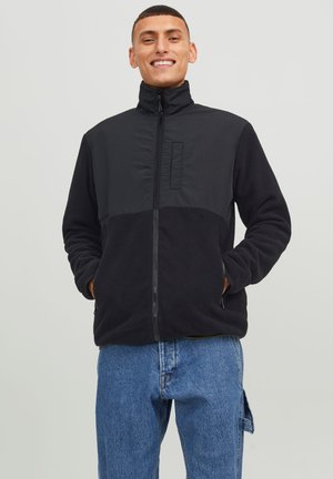 Black fleece jacket with a zip front, high collar, and contrasting fabric panel. Features side pockets and a relaxed fit. Denim jeans visible below.