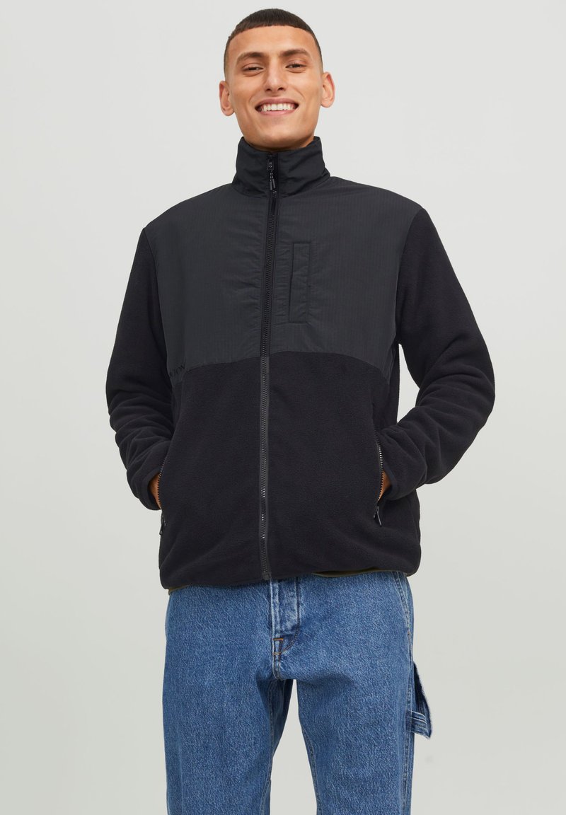 Svart fleecejakke med glidelås foran, høy krage og kontrastfarget stoffpanel. Har side lommer og en avslappet passform. Jeans av denim er synlige nedenfor.