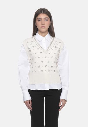 Maglione senza maniche lavorato a maglia crema con scollatura a V, impreziosito da borchie argentate, indossato sopra una camicia bianca con colletto, abbinato a pantaloni neri.