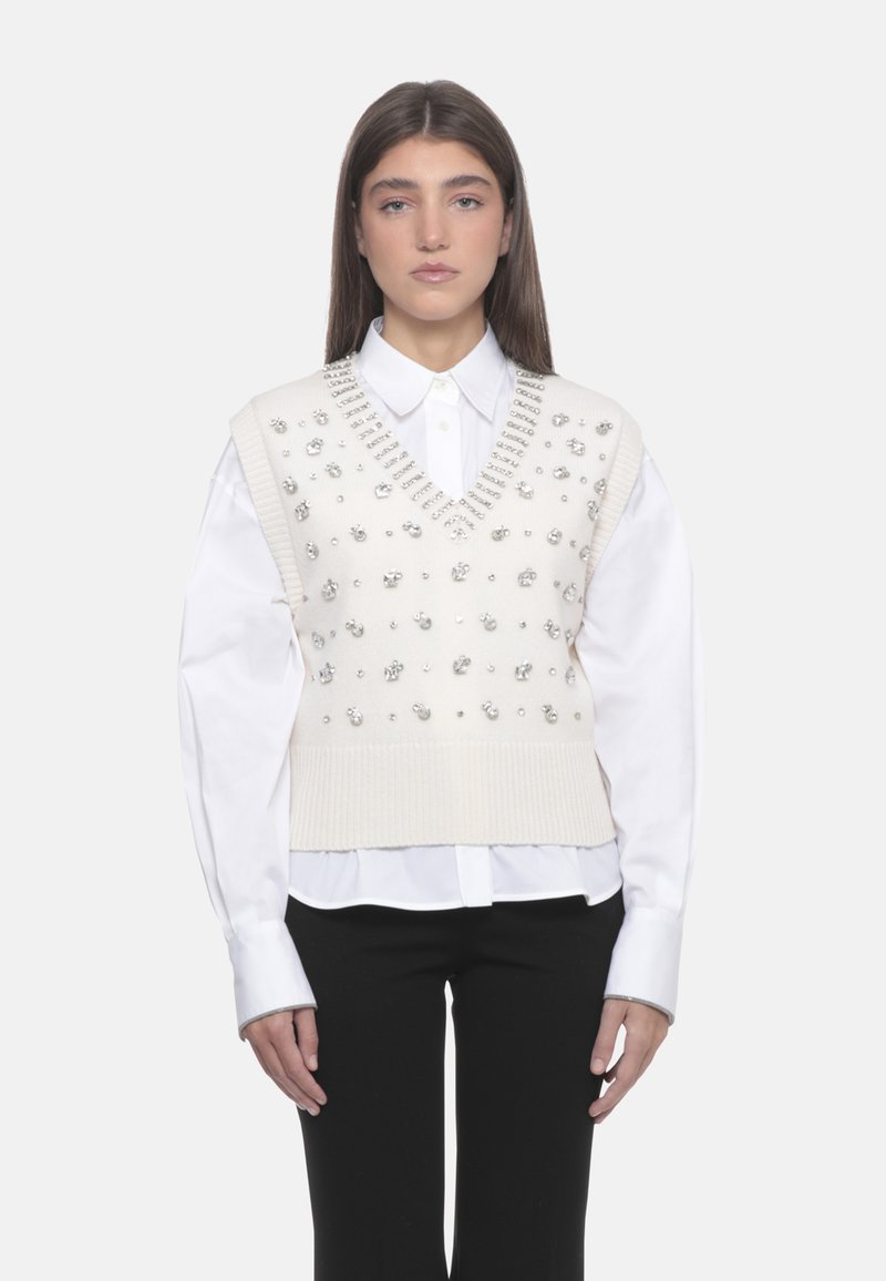 Maglione senza maniche lavorato a maglia crema con scollatura a V, impreziosito da borchie argentate, indossato sopra una camicia bianca con colletto, abbinato a pantaloni neri.