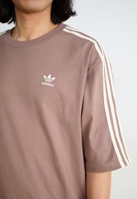 Person med en mauve Adidas T-shirt med tre vita ränder på axeln och Adidas-logotypen på bröstet, mot en enfärgad bakgrund.