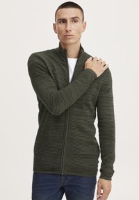 Mørkegrøn zip-up cardigan med vertikal ribstruktur, lange ærmer, høj krave og let figursyet design, parret med en hvid t-shirt underneath.
