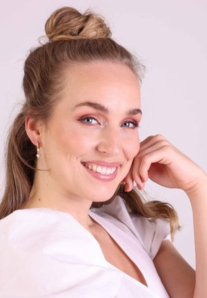 Portrait d'une personne avec de longs cheveux bruns ondulés rassemblés en chignon, portant des boucles d'oreilles en or avec des accents de perles et un chemisier blanc texturé.