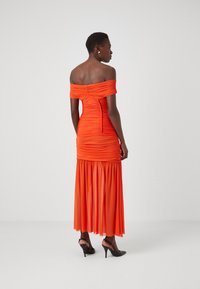 Φόρεμα πορτοκαλί off-the-shoulder με ριχτό ύφασμα, εφαρμοστή ζώνη και ρέουσα φούστα. Μαύρα ψηλοτάκουνα παπούτσια ολοκληρώνουν την εμφάνιση.