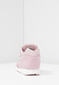 Nike-tenniskenkä pehmeässä vaaleanpunaisessa nahassa, jossa on valkoinen pohja ja teksturoituja yksityiskohtia. Mukana pehmustettu nilkkapanta ja kohokuvioitu logo kantapäässä.