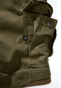 Alyvuogių žalios spalvos audinys su lygiu paviršiumi, kuriame yra kišenė su sagute ir mažas juodas etiketė. Pakraštyje yra rudos spalvos akcentas apačioje.