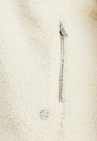 Beige textured fleece fabric with a small metallic zipper and a circular silver logo emblem below it.