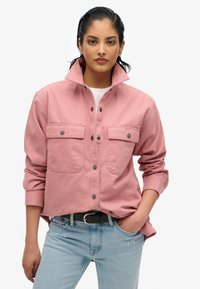 Chemise rose à boutons pression avec deux poches poitrine, boutons-pression, et col haut, associée à un jean bleu clair et une ceinture noire.
