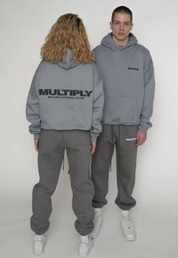 Sivá mikina s kapucňou s čiernym logom „MULTIPLY“ na chrbte, doplnená o zodpovedajúce sivé tepláky. Bavlnená zmes, voľný strih, s manžetami a vreckami.