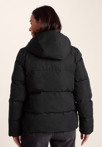 Woman wearing a black hooded puffer jacket and dark jeans, shown from the back against a plain light background.