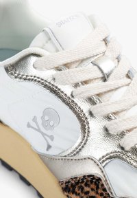Zapatillas metálicas plateadas con acentos de cuero blanco y un detalle de estampado de leopardo. Presenta un logo de calavera y huesos cruzados en el costado.
