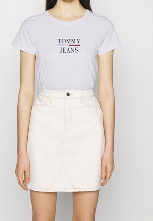 Nainen, joka pukeutuneena valkoiseen "Tommy Jeans" -t-paitaan ja valkoiseen korkeavyötäröiseen denimhameeseen, jossa on etutaskut ja musta nappi.
