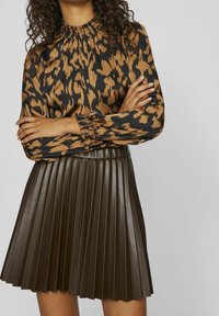Blusa marrone e nera a motivo con scollatura smock alta e maniche lunghe, abbinata a una gonna nera in ecopelle plissettata senza maniche.