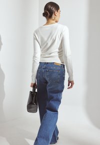 Valkoinen pitkähiha toppi ja siniset leveälahkeiset farkut. Musta käsilaukku kädessä. Yksinkertainen muotoilu, pehmeä tekstuuri, näkyvät saumat ja suuret takataskut.