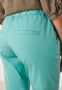 Turkosa byxor med elastisk midja som har en mjuk textur och bakfickor samt en tapered design. Tyget verkar lätt och flexibelt.